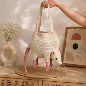 Op de afbeelding: Een crèmekleurige gehaakte tas in de vorm van een rat, voorzien van een lange roze staart, roze pootjes, roze oren, zwarte kraaloogjes en witte draadsnorharen. De tas heeft twee geïntegreerde gehaakte handvatten aan de bovenzijde.