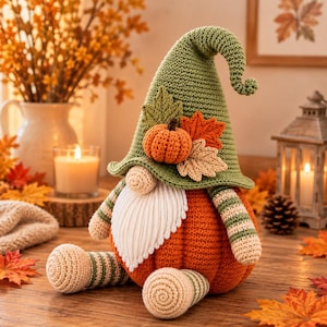 Könnte beinhalten: Eine gehäkelte Wichtelfigur mit einer hohen grünen Zipfelmütze, die mit einem kleinen orangefarbenen Kürbis und zwei Stoffblättern verziert ist. Der Wichtel hat eine runde beige Nase, einen weißen Garnbart und einen Körper wie ein Kürbis.