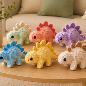 May include: Six small crocheted dinosaur plushies in pastel colors including light green, coral, purple, blue, yellow, and cream. Each dinosaur features a textured body, a row of soft spikes along the back, and two black bead eyes with pink cheeks.