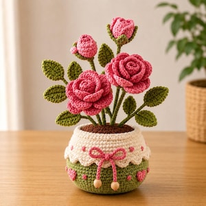 May include: A crocheted potted plant featuring three vibrant pink roses and two smaller buds with green leaves. The round pot is cream and green with a scalloped edge, decorated with small pink hearts and a pink yarn bow with two wooden beads.