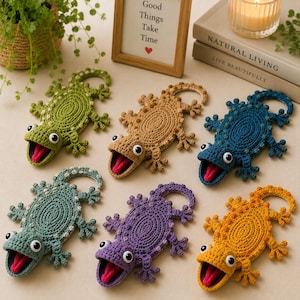 May include: Six crocheted lizard figures in green, tan, blue, teal, purple, and yellow. Each lizard features a textured body, a wide-open mouth with a protruding pink tongue, and two white and black eyes. The figures are arranged on a flat surface.