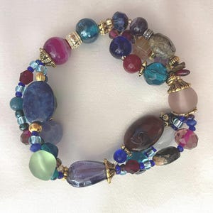Könnte beinhalten: Ein mehrsträngiges Armband mit einer Auswahl an Perlen in verschiedenen Formen, Größen und Farben, darunter Blau, Violett, Rosa, Grün und Braun. Das Design umfasst facettiertes Glas, glatte Steine und goldfarbene Metallzwischenperlen.