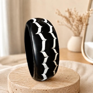 May include: A thick, rounded bangle bracelet featuring a glossy black finish with two vertical rows of repeating white, pointed, chevron-like patterns carved into the surface, creating a high-contrast graphic design on the circular band.