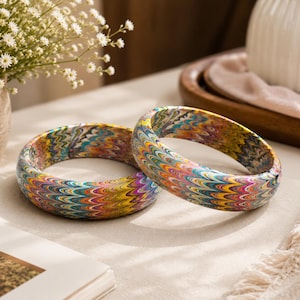 May include: Two circular bangles featuring a vibrant, multi-colored marbled pattern with wavy, repeating lines in shades of yellow, blue, red, and purple. The bracelets have a smooth, rounded exterior surface and a consistent, thick, curved profile.