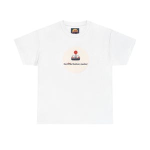 May include: A white short-sleeve t-shirt featuring a circular graphic on the chest. The graphic displays a black and red retro joystick icon above the text "Certified button masher" in a dark brown font. The shirt has a crew neckline.