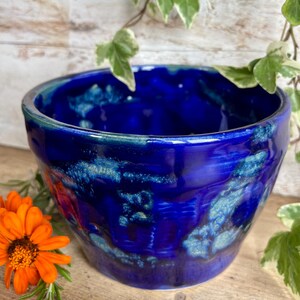 May include: A round ceramic bowl featuring a deep cobalt blue glaze with mottled turquoise and light blue accents. The surface has a glossy, reflective finish with visible variations in colour intensity across the exterior and interior walls.