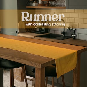May include: A mustard yellow table runner featuring horizontal contrasting stitching, laid flat across a wooden table. The text "Runner with contrasting stitching" is overlaid on the image, describing the textile product in a kitchen setting.