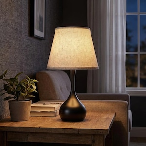 May include: A table lamp with a smooth, matte black teardrop-shaped base and a tapered, textured beige fabric lampshade. The lamp emits a warm, soft glow, illuminating the surrounding area while resting on a wooden surface.