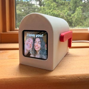 May include: A white, mailbox-shaped digital display device featuring a red flag on the side. The screen shows a photograph of two smiling people with the white text "I love you!" overlaid at the top. The device has a curved top and a square base.