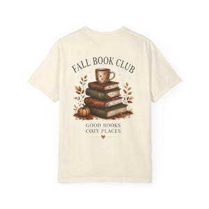 Op de afbeelding: Een crèmekleurig t-shirt met een afbeelding op de achterkant van een stapel boeken met een mok erop, omringd door herfstbladeren en een pompoen. De tekst luidt FALL BOOK CLUB en GOOD BOOKS COZY PLACES met een klein hartje eronder.