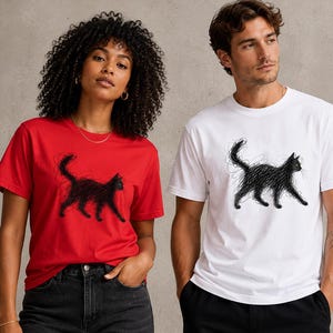 May include: A red t-shirt and a white t-shirt, each featuring a black graphic print of a cat drawn in a scribbled, sketchy style. The cat is depicted in profile, walking toward the right side of the shirt, with a raised tail and textured lines.