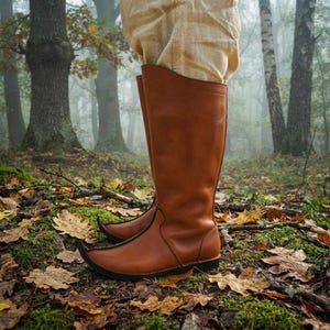 May include: A pair of knee-high brown leather boots with a pointed toe design and black trim along the sole. The boots feature a smooth finish and are worn with light tan fabric trousers that tuck into the top of the boot shafts.