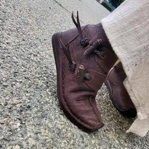 May include: A pair of dark brown leather ankle boots featuring a textured surface, a rounded toe, and a side closure system with thin leather laces and small wooden toggle fasteners. The boots have a flat sole and a visible seam along the edge.