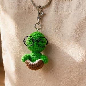 May include: A green crocheted turtle keychain featuring a round head with black wire-rimmed glasses and a brown shell with a white underside. The small stuffed figure is attached to a silver metal lobster clasp and a circular split ring.