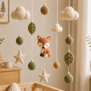 May include: A hanging nursery mobile featuring a central crocheted fox surrounded by dangling elements including white clouds, green leaves, textured spheres in shades of brown and green, and cream-coloured stars suspended from a circular frame.