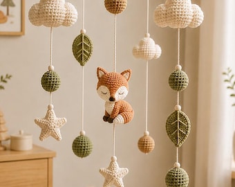 May include: A hanging nursery mobile featuring a central crocheted fox surrounded by dangling elements including white clouds, green leaves, textured spheres in shades of brown and green, and cream-colored stars suspended from a circular frame.