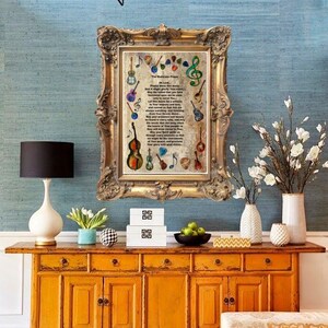 May include: A framed print with musical instruments and text hangs above a mustard yellow cabinet. The cabinet has multiple drawers and doors. A white lamp, vases, and decorative items sit on the cabinet. The wall is a light blue.