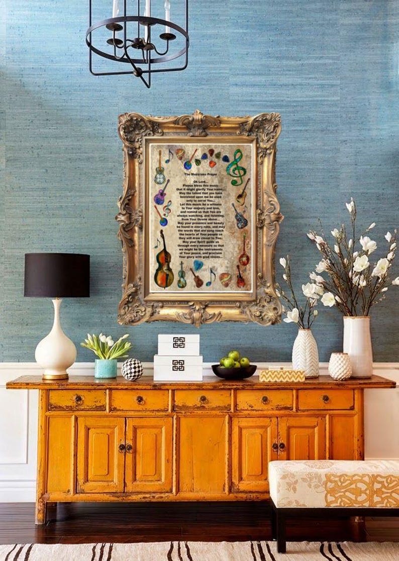 May include: A framed print with musical instruments and text hangs above a mustard yellow cabinet. The cabinet has multiple drawers and doors. A white lamp, vases, and decorative items sit on the cabinet. The wall is a light blue.