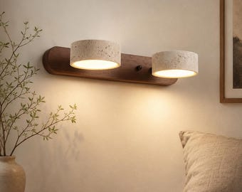 Può includere: Una lampada da parete con una base orizzontale in legno marrone scuro e due paralumi cilindrici in pietra beige porosa. Ogni paralume emette una luce calda rivolta verso il basso, illuminando la superficie della parete sotto la lampada.