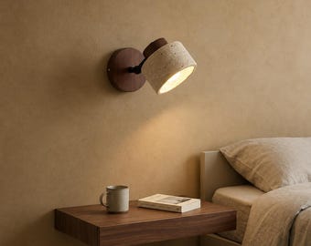 Può includere: Una lampada da parete con base circolare in legno scuro e paralume cilindrico in pietra beige porosa. L'apparecchio è orientabile e proietta una luce calda su un comodino sospeso in legno con sopra una tazza in ceramica e un libro.
