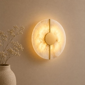 May include: A circular wall sconce featuring a translucent, marbled white stone disc illuminated from behind. A vertical gold metal bar bisects the disc, with a solid gold circular plate centered over the bar, creating a warm, glowing light effect.