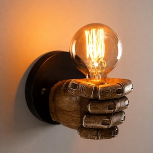 Puede incluir: Un aplique de pared que presenta una escultura de mano articulada de color marrón y textura rugosa, que sostiene una bombilla de cristal redonda y transparente con un filamento brillante visible. La mano se fija a una base circular negra.