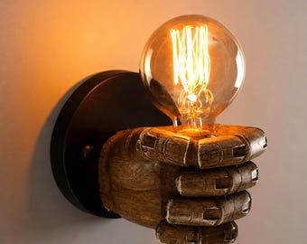 Può includere: Una lampada da parete con una scultura di mano articolata, di colore marrone e dalla superficie ruvida, che sostiene una lampadina in vetro rotonda e trasparente con un filamento luminoso visibile. La mano è fissata a una base circolare nera.