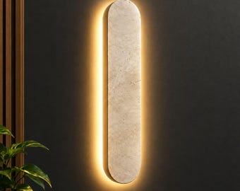 Può includere: Un'applique da parete verticale a forma di pillola con una superficie in pietra beige testurizzata. L'apparecchio emette una luce calda e soffusa dai bordi, creando un effetto alone sulla parete. Il design è allungato, liscio e minimalista.