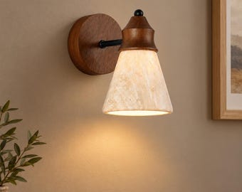 Può includere: Una lampada da parete con base circolare in legno scuro, braccio in metallo nero e paralume a forma di campana in pietra screziata color crema. La lampada emette una luce calda rivolta verso il basso, che mette in risalto la texture naturale.