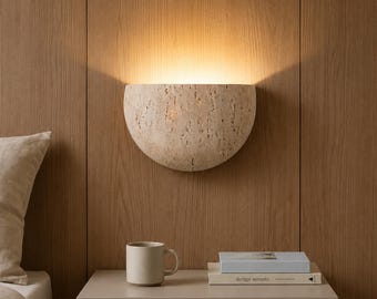 Può includere: Una lampada da parete semicircolare con una finitura in pietra beige porosa e testurizzata. L'apparecchio emette una luce calda rivolta verso l'alto contro la parete. La lampada è montata su una superficie in legno sopra un comodino.