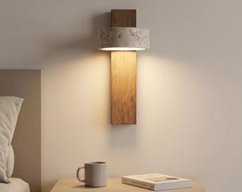 Può includere: Una lampada da parete con una piastra di supporto rettangolare in legno e un paralume cilindrico dalla texture grigia maculata simile alla pietra. La lampada emette una luce calda rivolta verso il basso, illuminando la parete sottostante.