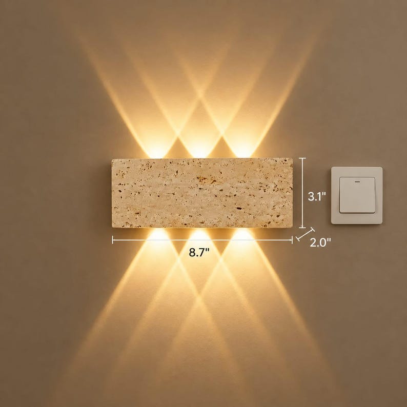 May include: A rectangular beige stone wall lamp with a porous texture, emitting warm light beams upward and downward. The fixture measures 8.7 inches in width, 3.1 inches in height, and 2.0 inches in depth, positioned next to a white light switch.