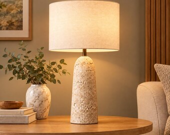 Può includere: Una lampada da tavolo con una base cilindrica in pietra beige testurizzata dalla superficie porosa e un paralume a tamburo in tessuto bianco sporco. La lampada emette una luce calda e soffusa, illuminando l'area circostante con delicatezza.