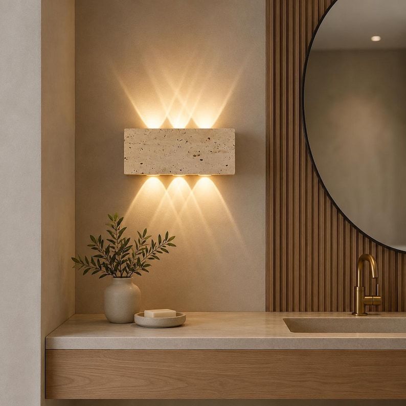 May include: A rectangular wall sconce made of beige porous stone with a textured surface. The fixture emits warm light in three distinct beams directed both upward and downward, creating a symmetrical pattern of illumination on the wall surface.