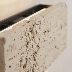 May include: A rectangular wall light fixture featuring a thick, porous beige travertine stone front panel with a natural, uneven texture. The stone is mounted on a dark metal frame that emits a warm, soft glow from behind the edges of the stone.