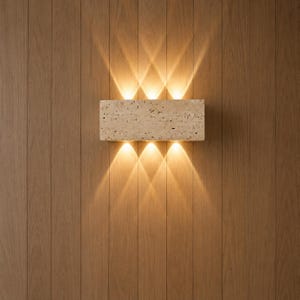 May include: A rectangular wall sconce made of beige porous stone with a textured surface. The fixture emits light in three distinct upward beams and three distinct downward beams, creating a symmetrical pattern of warm illumination on the wall.