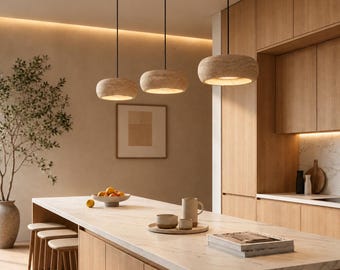 May include: Three circular pendant lights with a textured beige stone finish hang from thin black cords above a kitchen island. The lights feature a hollow center that emits a warm glow, illuminating the marble countertop and wooden cabinetry below.