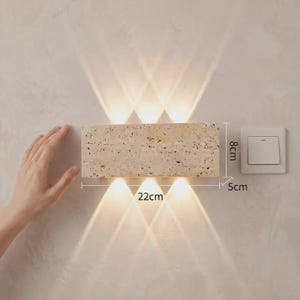 May include: A rectangular beige travertine wall lamp with a porous texture, emitting light in six distinct beams upward and downward. The fixture measures 22 cm in length, 8 cm in height, and 5 cm in depth, positioned next to a white light switch.