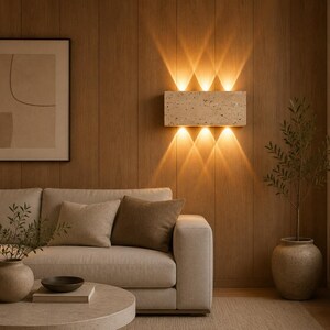 May include: A rectangular wall-mounted light fixture made of beige porous stone. The fixture features three upward-facing and three downward-facing light beams that create a symmetrical pattern of warm illumination against the wooden wall surface.