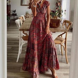 May include: A floor-length maxi dress featuring a red, orange, and brown paisley pattern. The garment has short puff sleeves, a smocked bodice, and a tiered skirt. The fabric displays a detailed, intricate print throughout the entire piece.