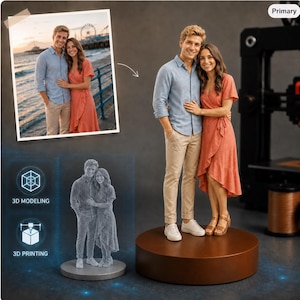 Puede incluir: Una figura impresa en 3D personalizada de una pareja, basada en una fotografía. La figura muestra a una persona con camisa azul claro y pantalones beige, y a otra con un vestido cruzado color coral, sobre una base circular.