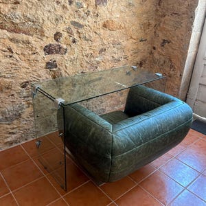 Puede incluir: Un escritorio de cristal con una superficie rectangular transparente y paneles laterales unidos por soportes de esquina de metal plateado, situado sobre un sillón bajo y redondeado tapizado en cuero verde salvia desgastado con costuras horizontales.