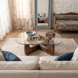May include: A coffee table with a clear glass top and a dark brown wooden base featuring an intersecting design. The table holds a blue and white patterned bowl, a small wooden bird figurine, and a brass tray with green glass stones.