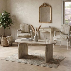 Puede incluir: Una mesa de centro ovalada de madera clara con dos patas de panel curvado sobre una alfombra estampada. La habitación cuenta con dos sillones tapizados, una mesa consola con un espejo de marco dorado, una planta y varios libros.