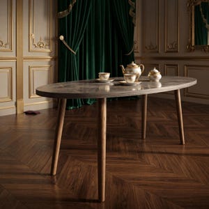 Puede incluir: Una mesa de comedor ovalada con una superficie de patrón de mármol gris y cuatro patas de madera marrón claro. Sobre la mesa hay una tetera de porcelana blanca y dorada, un azucarero a juego y dos tazas blancas y doradas con platillos.