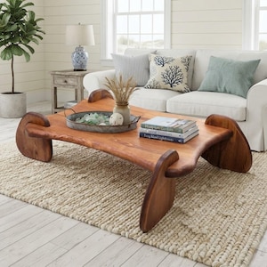 May include: A low-profile coffee table crafted from natural wood with a smooth, curved surface and four distinct, rounded slab legs. The table features a prominent grain pattern and a warm, medium-brown finish, providing a sturdy base for decor.