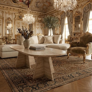 Peut inclure: Une table basse ovale en travertin avec un pied central repose sur un tapis à motifs. La table supporte un vase en céramique avec du feuillage séché et une pile de livres. La pièce présente des moulures dorées, des lustres et des sièges.