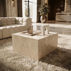 Puede incluir: Una mesa de centro cuadrada de color beige con textura de piedra y superficie lisa. Sobre la mesa hay una bandeja rectangular, un vaso de cristal transparente y un pequeño objeto decorativo blanco en forma de arco sobre una alfombra beige.