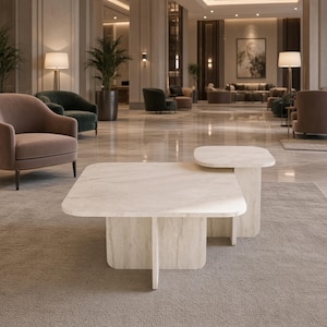 Op de afbeelding: Een salontafel met twee niveaus gemaakt van lichtbeige steen met een zichtbaar natuurlijk aderpatroon. De tafel heeft een groot vierkant onderoppervlak en een kleiner verhoogd vierkant oppervlak, beide ondersteund door kruisende verticale plaatpoten.
