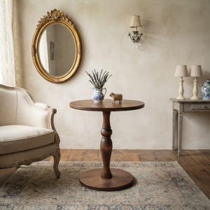 May include: A round wooden pedestal table with a dark brown finish, featuring a central turned column base and a circular top. The table is positioned in a room with a beige armchair, a gold oval mirror, and a small wooden sheep figurine.